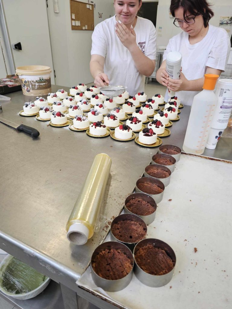 Két fehér inget viselő ember számos kis tortát díszít, tetején tejszínhabbal és bogyós gyümölcsökkel egy nagy fémasztalon. A közelben kerek csokoládés sütemények vannak fémformákban, és különféle sütőeszközök és hozzávalók vannak szétszórva.