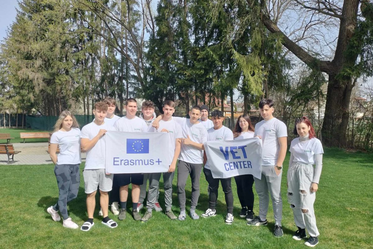Egy tízfős, fehér pólót viselő fiatal felnőtt csoport áll a szabadban egy füves területen, háttérben fákkal. Két transzparenst tartanak a kezükben: az egyiken „Erasmus+” felirat olvasható EU-zászlóval, a másikon „VET Center”. Úgy tűnik, hogy részt vesznek egy eseményen vagy programon.