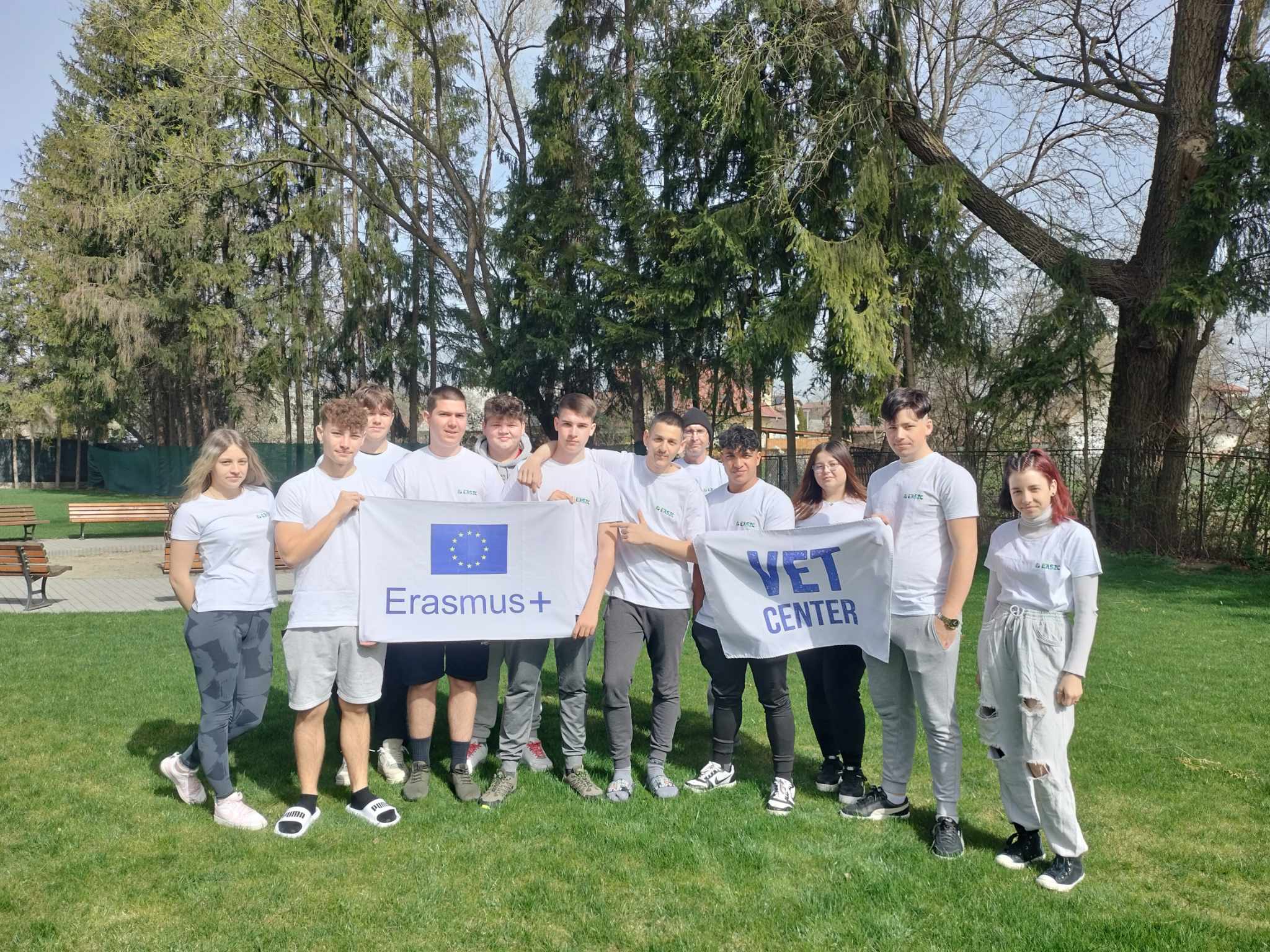 Egy tízfős, fehér pólót viselő fiatal felnőtt csoport áll a szabadban egy füves területen, háttérben fákkal. Két transzparenst tartanak a kezükben: az egyiken „Erasmus+” felirat olvasható EU-zászlóval, a másikon „VET Center”. Úgy tűnik, hogy részt vesznek egy eseményen vagy programon.