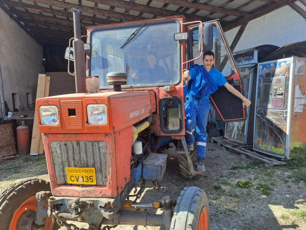 Egy fedett területen parkoló narancssárga-piros mezőgazdasági traktor vezetőfülkéjéből egy kék munkaruhát viselő személy lép ki. Egy másik személy látható a traktor fülkéjében. Különféle felszerelések és eszközök vannak szétszórva a területen.
