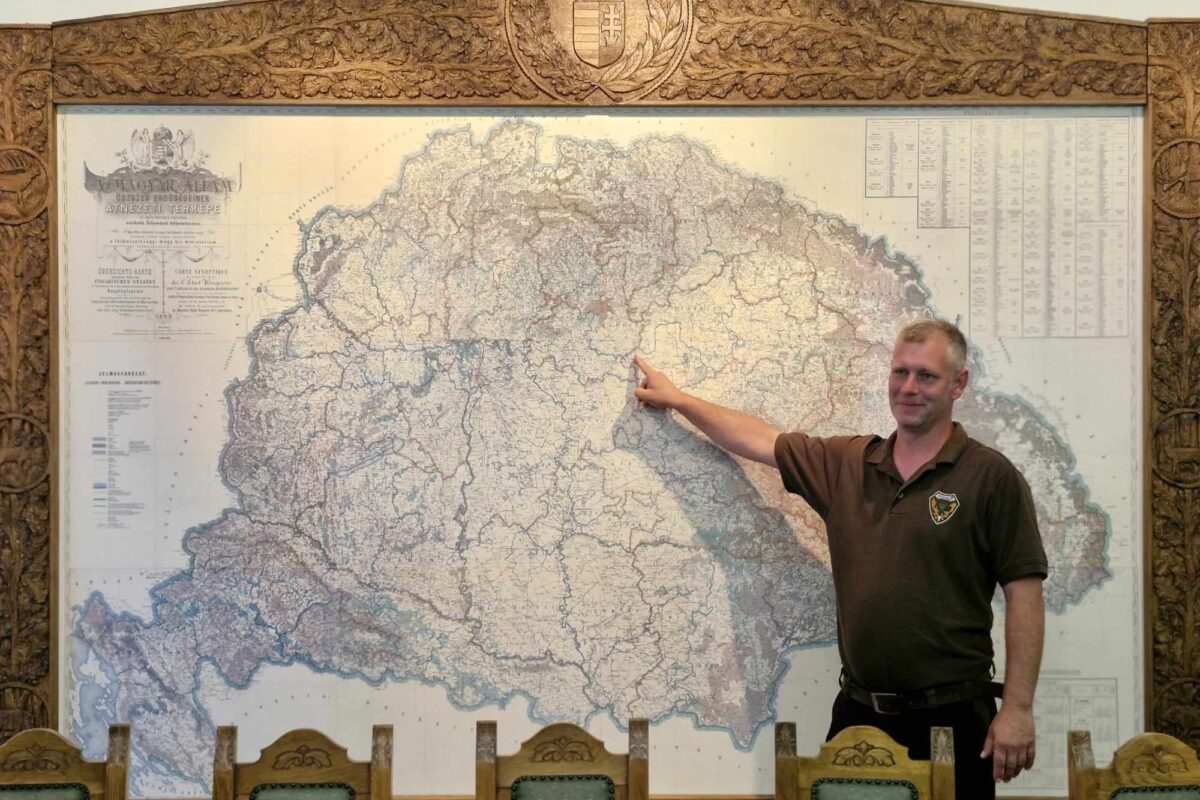 Egy barna inget viselő férfi mutat Magyarország nagy, bonyolult térképére, amelyet díszes fafaragványok kereteznek. Egy sor díszes háttámlájú faszék mellett áll. A részletes térkép történelminek tűnik, különféle földrajzi és politikai jelölésekkel.