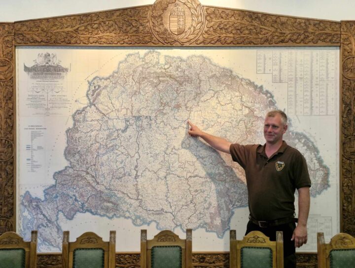 Egy barna inget viselő férfi mutat Magyarország nagy, bonyolult térképére, amelyet díszes fafaragványok kereteznek. Egy sor díszes háttámlájú faszék mellett áll. A részletes térkép történelminek tűnik, különféle földrajzi és politikai jelölésekkel.