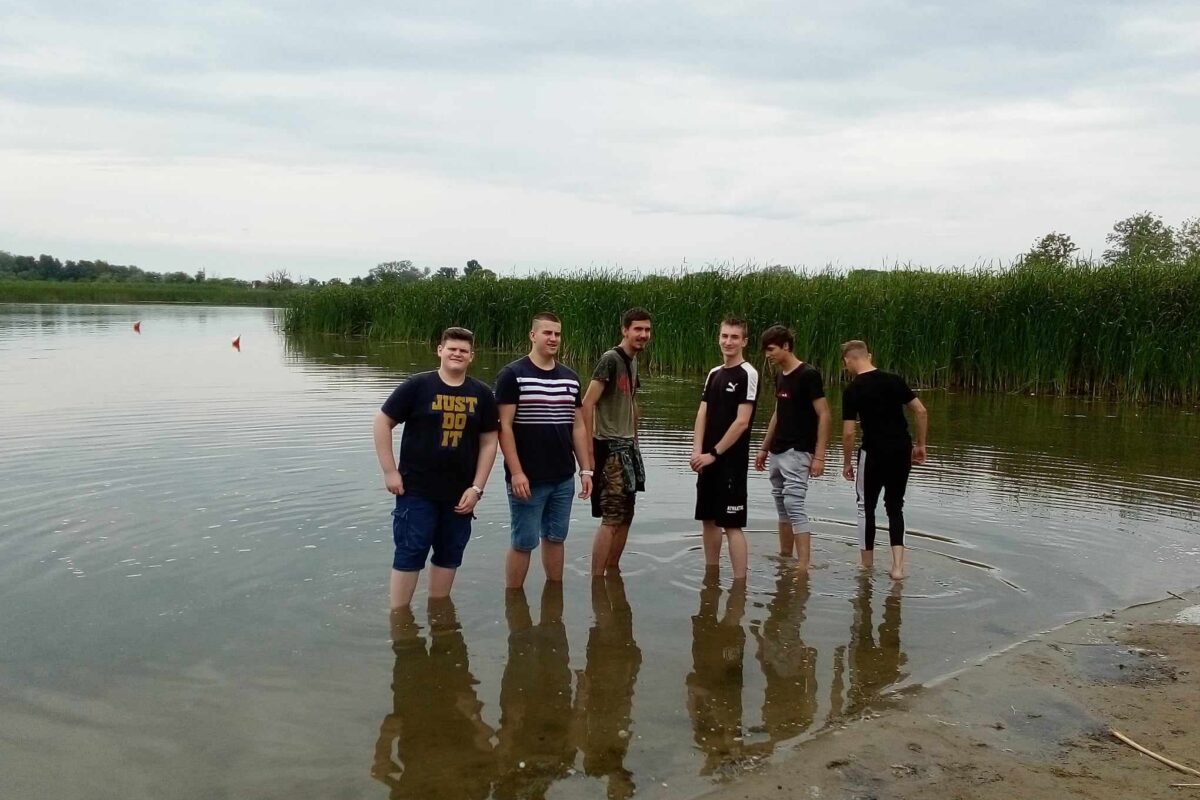 Egy hat fiatal férfiból álló csoport sekély vízben áll egy tó partján, részben borjaikig elmerülve. A háttérben magas nád és felhős égbolt látható, ami borús napra utal. Úgy tűnik, hogy egy laza pillanatot élveznek a víz mellett.