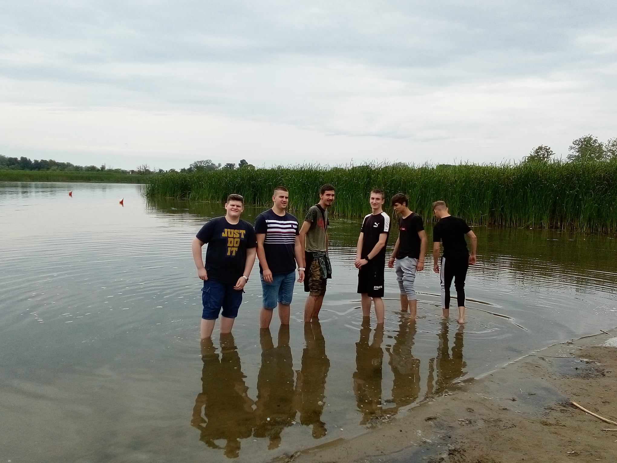 Egy hat fiatal férfiból álló csoport sekély vízben áll egy tó partján, részben borjaikig elmerülve. A háttérben magas nád és felhős égbolt látható, ami borús napra utal. Úgy tűnik, hogy egy laza pillanatot élveznek a víz mellett.