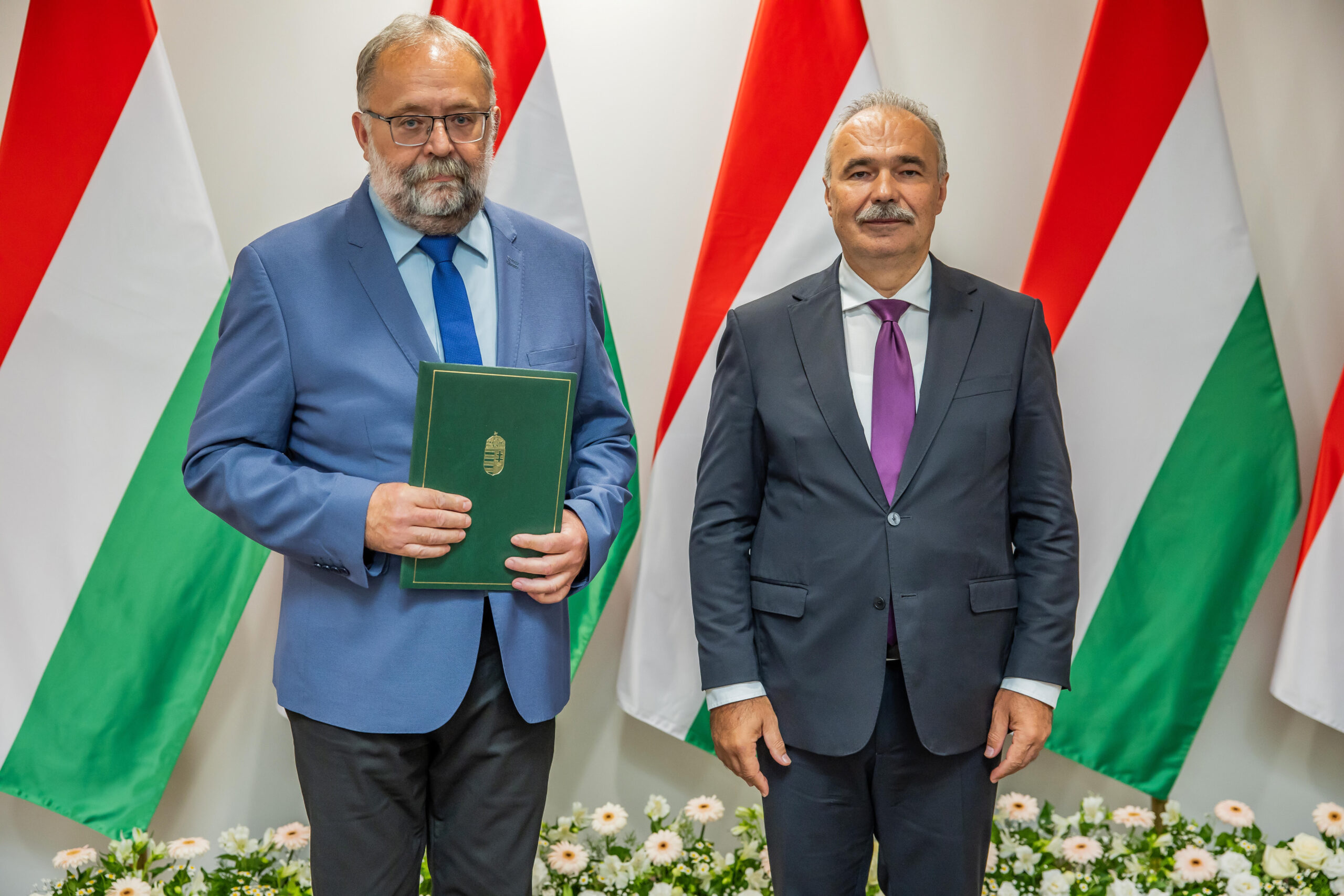 Két férfi áll egymás mellett a magyar zászlókat ábrázoló háttér előtt. A bal oldali férfi egy zöld, arany emblémával ellátott mappát tart, míg a jobb oldali férfi az oldala mellett tartja a kezét. Mindketten öltönyt viselnek; a bal férfi kék nyakkendőt, a jobb lila nyakkendőt visel.