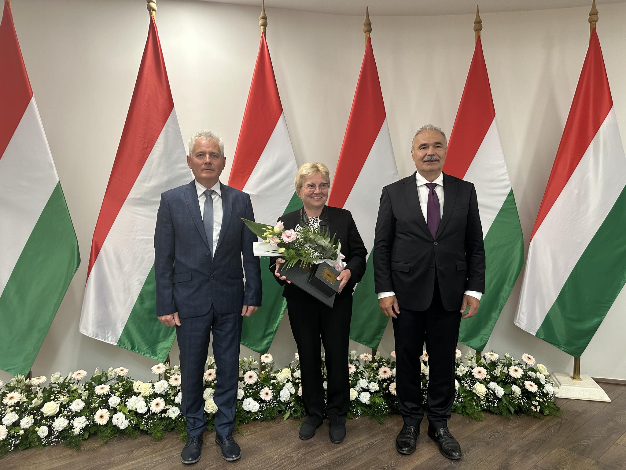 Több magyar zászló előtt hárman állnak sorban. A középső, virágcsokorral és kitüntetéssel tartó személy mellett két öltönyös férfi áll. A háttérben a padló mentén virágdíszek láthatók.